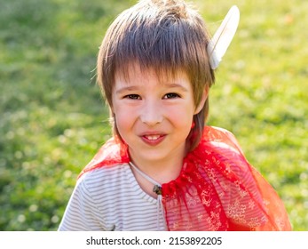 Portrait Of Smiling Boy Playing American Indian. Kid With White Bird Feather And Red Cloak. Costume Role Play. Outdoor Leisure Activity.