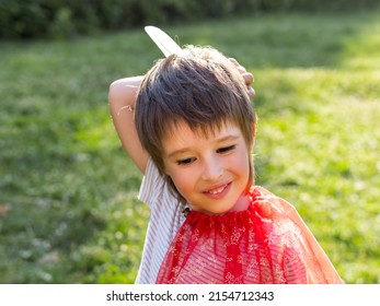 Portrait Of Smiling Boy Playing American Indian. Kid With White Bird Feather And Red Cloak. Costume Role Play. Outdoor Leisure Activity.