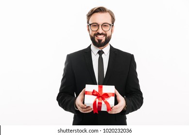 Portrait Of Smiling Brunette Man In Black Suit Looking On Camera And Holding Small Gift Box In Hands Isolated Over White Background