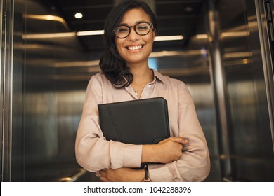 Portrait of smiling business woman holding a file standing in office elevator. Happy female executive looking at camera and smiling.