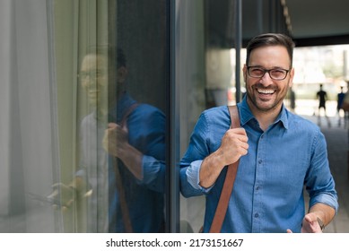 Portrait Of Smiling Businessman Carrying Laptop Bag. Young Male Professional Is Wearing Formals. He Is Standing By Window Of Office Building In The City.