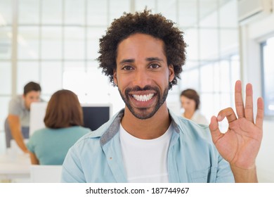 Portrait of a smiling businessman gesturing ok sign with colleagues in meeting in background at the office - Powered by Shutterstock