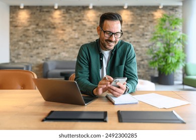 Portrait of smiling businessman reading messages over mobile phone while working over laptop at desk - Powered by Shutterstock