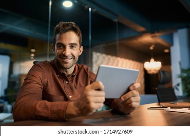 Portrait Of A Smiling Businessman Sitting After Hours Alone At His Dark Office Desk And Working Online With A Digital Tablet