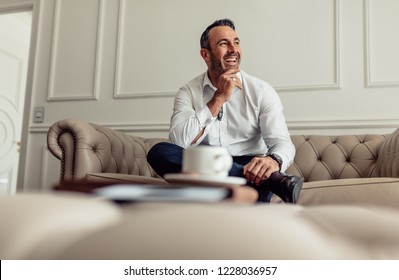 Portrait of smiling businessman sitting on sofa in hotel room and looking away. Mature businessman staying in luxurious hotel room on business trip.