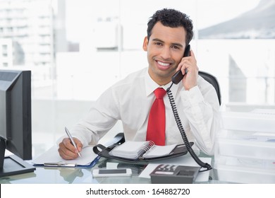 Portrait Of A Smiling Businessman Writing Notes While Using Land Line Phone At A Bright Office