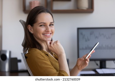 Portrait Of Smiling Businesswoman Sit On Ergonomics Chair Holds Smartphone, Take Break At Workplace Desk With Pc Look At Camera, Sales Stats On Computer Monitor. Tech, Business Apps, Analytics Concept