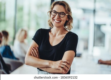 Portrait of smiling businesswoman standing in office with colleagues meeting in background. Successful female professional with her arms crossed in meeting room.