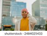 Portrait of a smiling cancer survivor in her 30s wearing a pink headscarf and yellow sweater, exuding confidence in an urban setting.