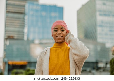 Portrait of a smiling cancer survivor in her 30s wearing a pink headscarf and yellow sweater, exuding confidence in an urban setting. - Powered by Shutterstock