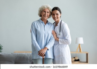 Portrait Smiling Caregiver And Older Woman With Walking Cane Standing At Home, Caring Doctor Wearing White Uniform Coat Hugging, Supporting Mature Female Patient, Looking At Camera, Healthcare