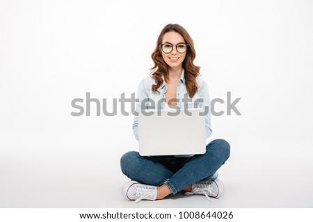Portrait of a smiling casual girl holding laptop computer while sitting on a floor isolated over white background