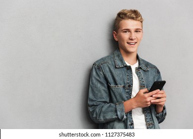 Portrait of a smiling casual teenage boy dressed in denim jacket holding mobile phone isolated over gray background - Powered by Shutterstock