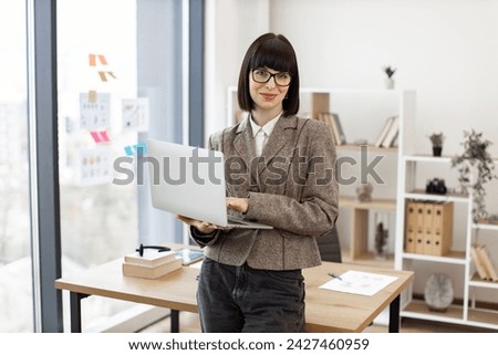 Portrait of smiling caucasian adult businesswoman sitting on edge of wooden desk with laptop. Cheerful woman in formal wear managing successful company from modern workplace.