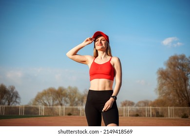 Portrait Of Smiling Caucasian Athletic Woman In Red Cap, Red Top And Black Cycling Shorts Outdoors At The Sports Ground In Spring.