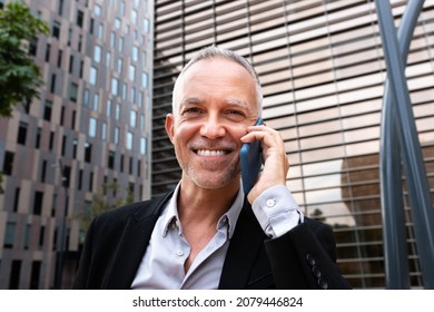 Portrait Of Smiling Caucasian Business Man Talking On Cell Phone Looking At Camera Next To Office Buildings.