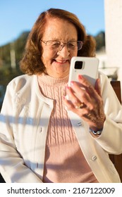 Portrait Of Smiling Caucasian Older Woman Making A Video Call Through Mobile Phone. She Is Outdoors.