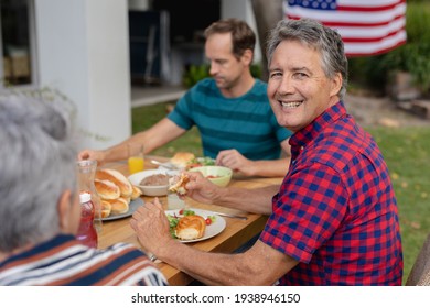 Portrait Of Smiling Caucasian Senior Man Sitting At Table With Family Having Meal In Garden. Three Generation Family Celebrating Independence Day Eating Outdoors Together.