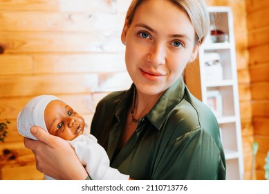 Portrait Of Smiling Caucasian Young Woman Holding Newborn African American Baby Doll In Her Arms And Looking At Camera, Indoors. Toy Child Reborn Handmade.