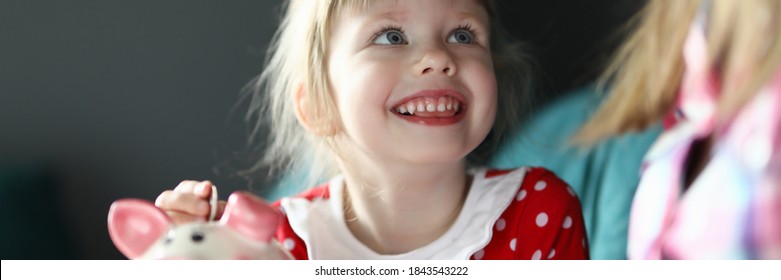 Portrait Of Smiling Cheerful Kid Putting Coin In Piggy Bank And Looking At Mother. Happy Joyful Child Wearing Red Dress In Dots. Investment And Finance Concept