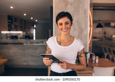 Portrait of a smiling coffee shop owner standing inside her shop. Female restaurant owner standing with a digital tablet.