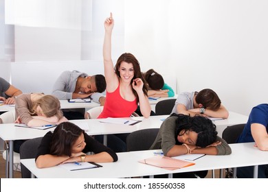 Portrait Of Smiling College Student Raising Hand With Classmates Sleeping At Desk In Classroom