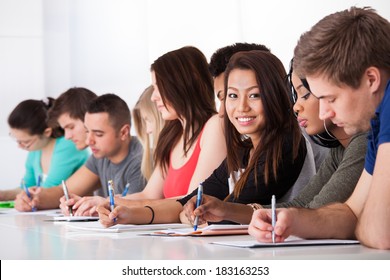 Portrait Of Smiling College Student Sitting With Classmates Writing At Desk