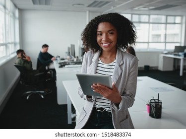 Portrait of a smiling confident african american young businesswoman holding digital tablet in hand looking at camera with colleague at background in office