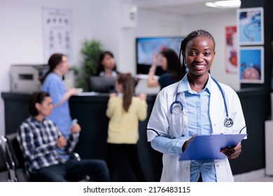 Portrait Of Smiling Confident Doctor In Busy Private Hospital Waiting For Next Appointment Holding Clipboard. Friendly Medic In White Lab Coat With Stethoscope Standing In Clinic Waiting Room.