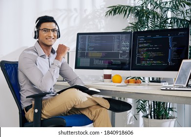 Portrait of smiling confident Indian coder sitting at his office desk with programming code on computer screens