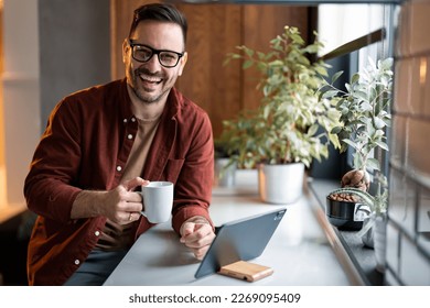 Portrait of smiling confident man looking at camera sitting indoors at home, holding a cup of coffee, using digital tablet technology device. Happy freelance worker looking at camera sitting at home. - Powered by Shutterstock