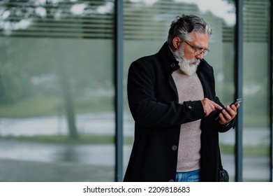 Portrait Of A Smiling And Confident Mature Businessman Walking Outside While Going To A Meeting And Using A Smartphone. Senior Bearded Man Dressed In Stylish Black Coat Outside Of The Office Building.