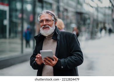 Portrait Of A Smiling And Confident Mature Businessman Walking Outside While Going To A Meeting And Using A Tablet. Senior Bearded Man Dressed In Stylish Black Coat Using Technology And Internet.