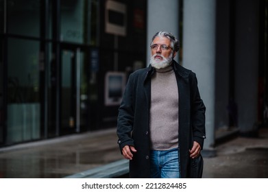 Portrait Of A Smiling And Confident Mature Businessman Walking Outside While Going To A Meeting. Senior Bearded Man Dressed In Stylish Black Coat Outside Of The Office Building Copy Space. .