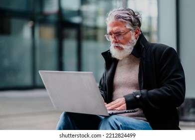 Portrait Of A Smiling And Confident Mature Businessman Sitting On The Bench Outside And Using Laptop. Senior Bearded Man Dressed In Black Coat Typing On A Computer Outside Of The Office Building.