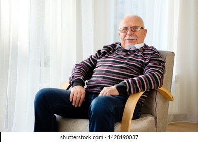 Portrait Of Smiling And Confident Senior Man 70-75 Years Old With Eyeglasses Relaxing In Armchair Near Light Window At Home. Elder Male Looking At Camera