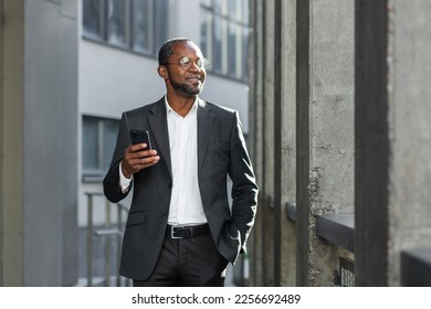 Portrait of a smiling confident and successful African American businessman in a suit and glasses standing outside near an office center, holding a phone in his hand. He keeps his hand in his pocket, - Shutterstock ID 2256692489