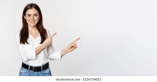 Portrait of smiling confident woman, business girl showing advertisement, pointing fingers right at logo, banner or announcement, standing over white background. - Powered by Shutterstock