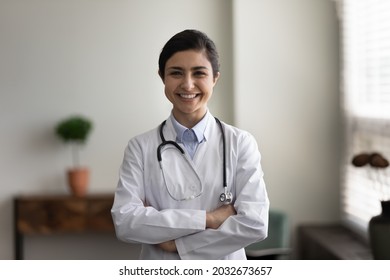 Portrait Of Smiling Confident Young Indian Ethnicity Female Gp Doctor Therapist In White Medical Coat Standing In Modern Office Room With Folded Hands, Professional Guardianship, Healthcare Concept.