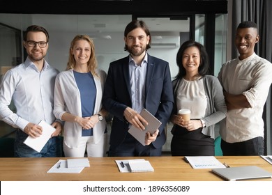 Portrait of smiling confident young and middle aged diverse office workers standing near table. Happy skilled caucasian male team leader posing for photo with mixed race colleagues at workplace.