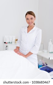 portrait of smiling cosmetologist  in white coat standing at empty massage table in salon