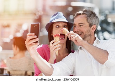 Portrait of a smiling couple eating ice cream and having fun in the city. The grey hair man with a beard is taking a selfie with phone. they make funny faces - Powered by Shutterstock