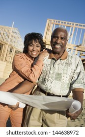 Portrait Of A Smiling Couple Holding Blueprint In Front Of House Construction Site