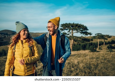 Portrait of a smiling couple of tourists dressed in warm clothes walking on a grassy mountain ridge during a sunset. Young people hiking in nature. Man hugging woman while having fun in the nature. - Powered by Shutterstock