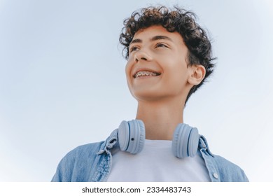 Portrait of smiling curly haired teenager with braces wearing headphones looking away standing on the street. Inspiration, technology concept