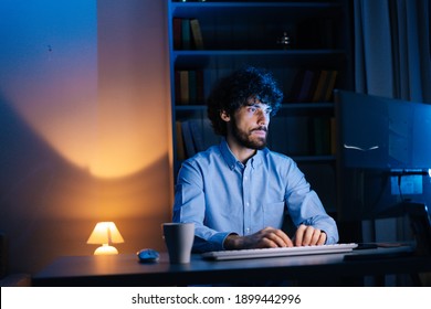 Portrait of smiling curly man looking intently at monitor screen while working on computer at late night. Hacker at work stealing accounts database. Concept of remote working. - Powered by Shutterstock