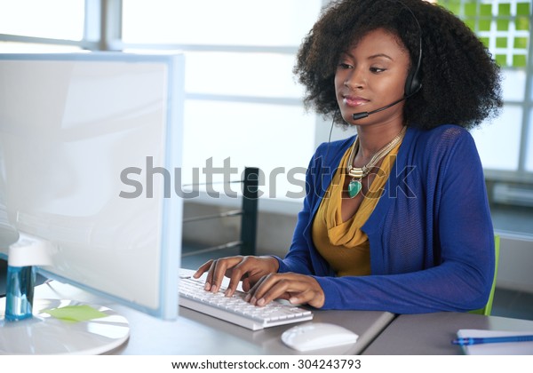 Portrait of a smiling customer\
service representative with an afro at the computer using\
headset