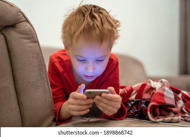 Portrait of smiling cute child lies on sofa and plays cheerfully in a game off phone. Gadget addiction. Mobile addict concept. - Powered by Shutterstock