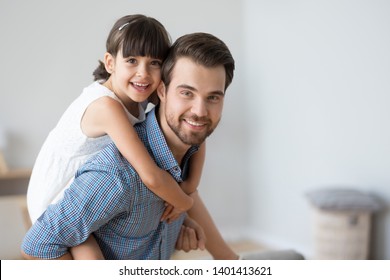 Portrait of smiling cute little preschooler girl piggyback young father looking at camera, loving dad carry funny small daughter on back, posing for picture together, parent and child hug make photo