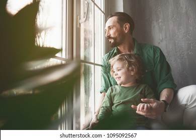 Portrait Of Smiling Dad And Outgoing Kid Looking At Window While Locating In Apartment